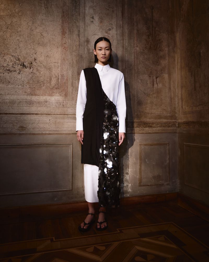 Elegant Asian woman in a white shirt dress with a black draped accent, standing in a room with warm, textured walls and parquet flooring.