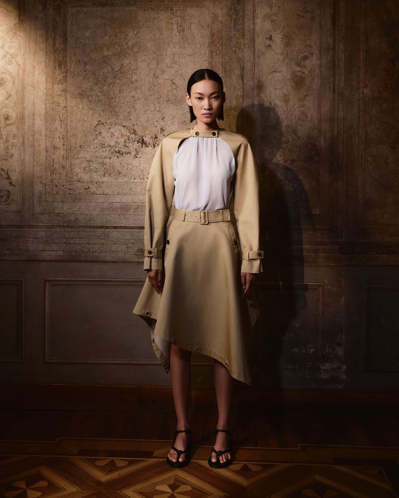 Fashion shot of an Asian woman in a tan belted trench coat dress and black sandals, posing against a distressed wall in a studio.