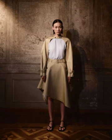 Fashion shot of an Asian woman in a tan belted trench coat dress and black sandals, posing against a distressed wall in a studio.