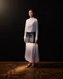 Model standing in a white pinstripe shirt and skirt with a black leather overlay panel against a dark background.