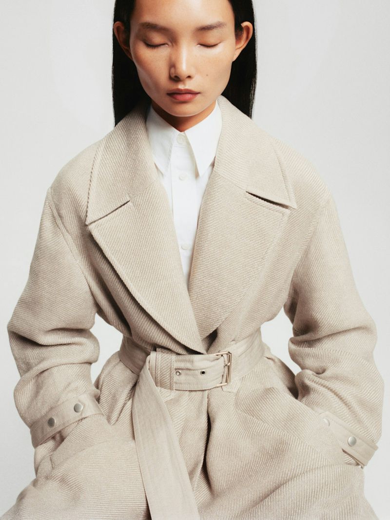 Close-up of an adult female model with eyes closed wearing a beige wool trench coat and white collared shirt against a white background.