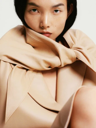 Close-up portrait of an adult woman with dark hair wearing a beige leather coat, set against a plain background with diffused lighting.
