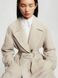 Ling Chen wearing an oversized belted beige coat over a crisp white collared shirt with eyes closed against a grey studio backdrop.