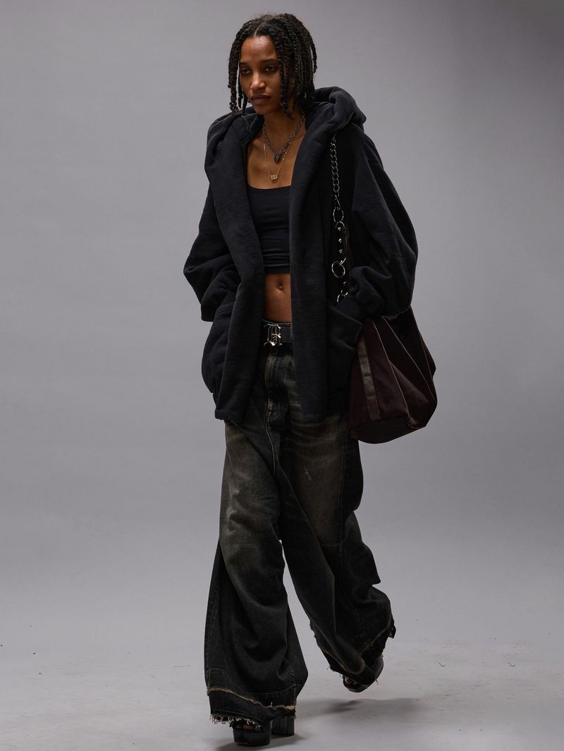 Young adult female fashion model in a hooded jacket, tank top, and wide-leg jeans poses in a studio. She carries a tote bag.