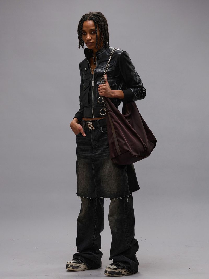 Full-length studio shot of an adult female with braided hair wearing a black leather jacket, distressed denim skirt and jeans, holding a brown bag.