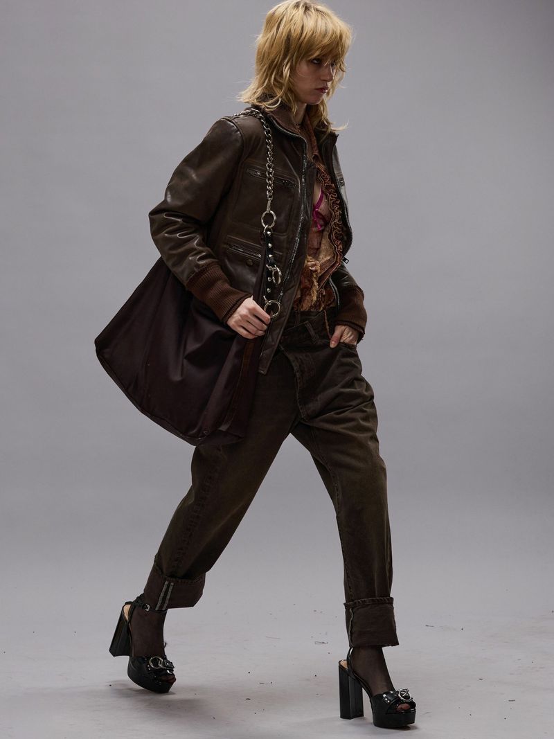 A woman walks in a studio wearing a brown jacket, matching pants, and black platform heels, carrying a brown bag, against a gray backdrop.