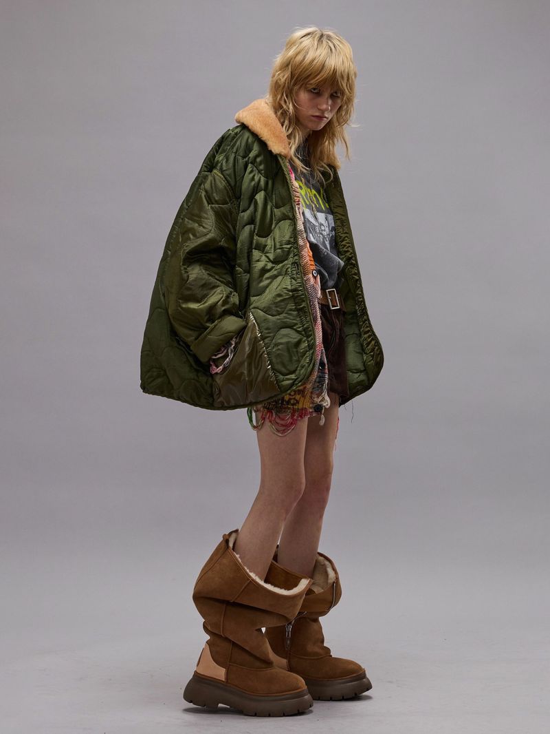 Woman with long blonde hair, wearing an olive drab coat with a fur collar, printed top and shorts, and brown boots poses in studio.