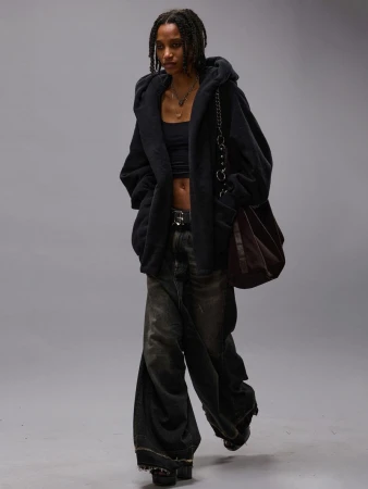 Model wearing a black hoodie over a black crop top and dark wide-leg jeans with a large shoulder bag against a grey seamless background