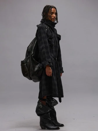 Model in a dark plaid coat, slouchy black leather boots, and large black backpack standing against a neutral grey studio backdrop.