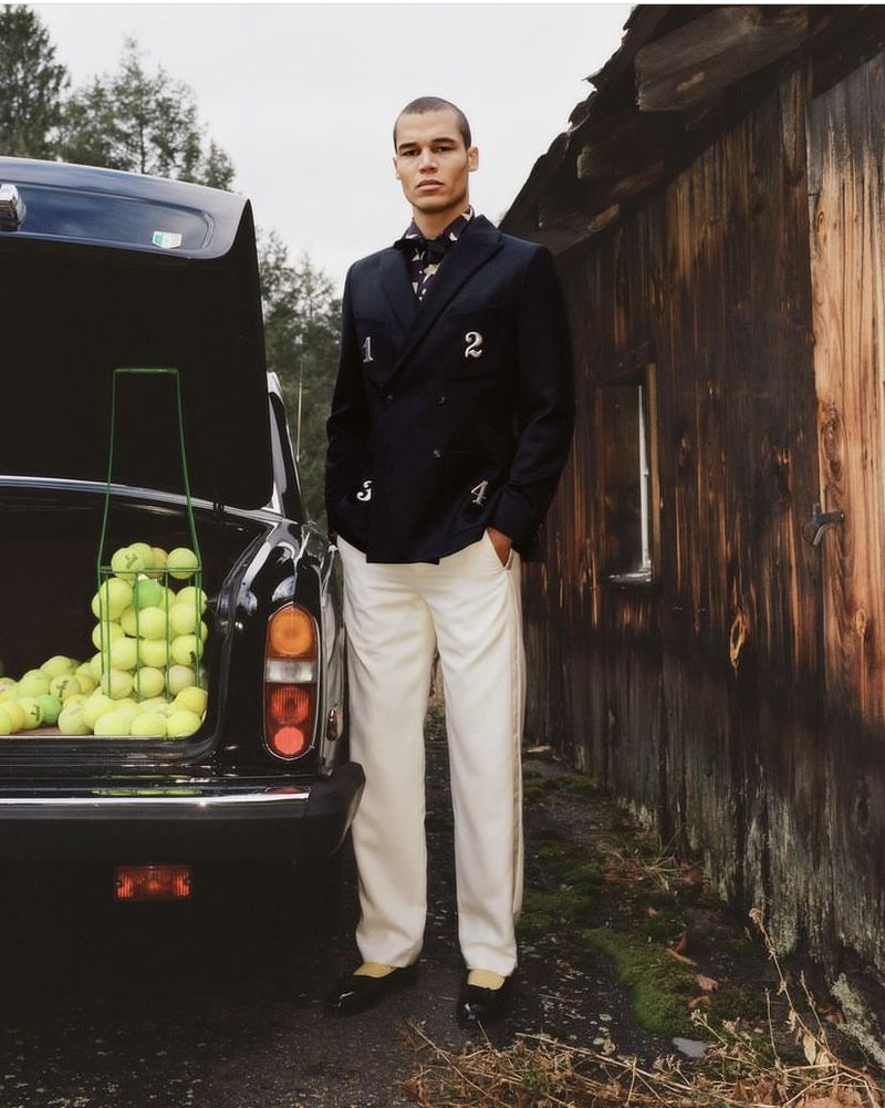 A well-dressed man in a blazer stands next to an old car with its trunk open, revealing a carrier full of tennis balls in a rustic outdoor setting.