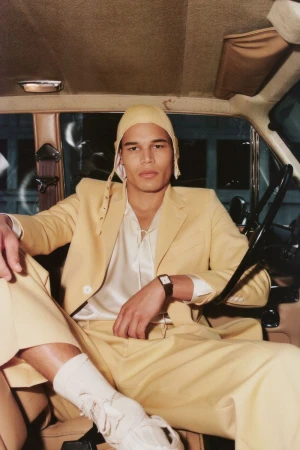 Model seated inside a car wearing a yellow suit, matching cap, and white socks for Bode fall winter 24 collection
