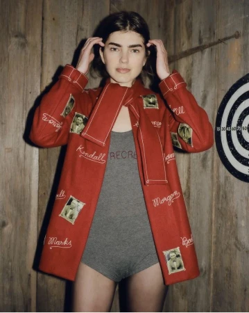 Woman in a red embroidered jacket poses indoors against a wood paneled wall with a dart board in the background in a medium shot.