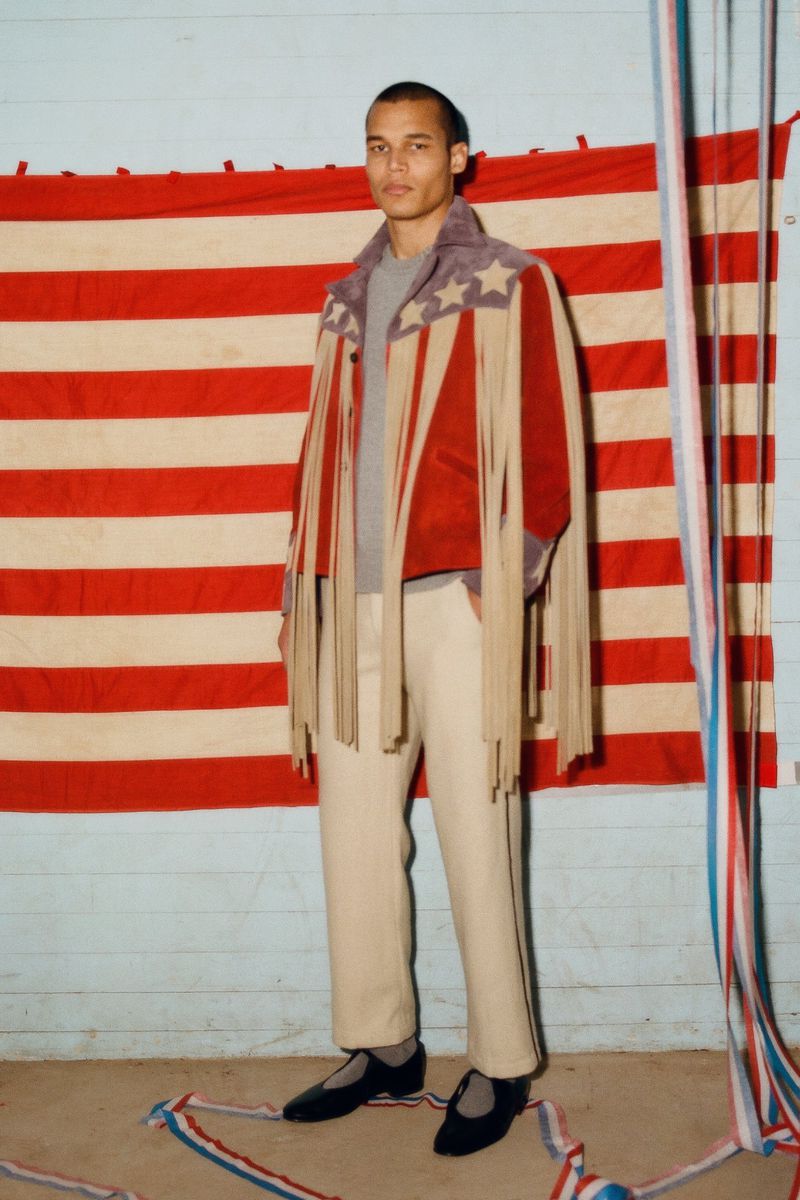 Man wearing a fringed stars and stripes jacket and cream trousers stands in front of an American flag. Fashion editorial.