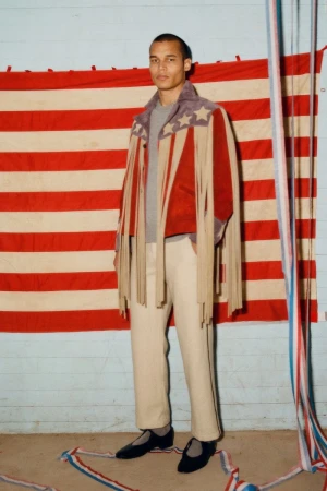 Man wearing a fringed stars and stripes jacket and cream trousers stands in front of an American flag. Fashion editorial.