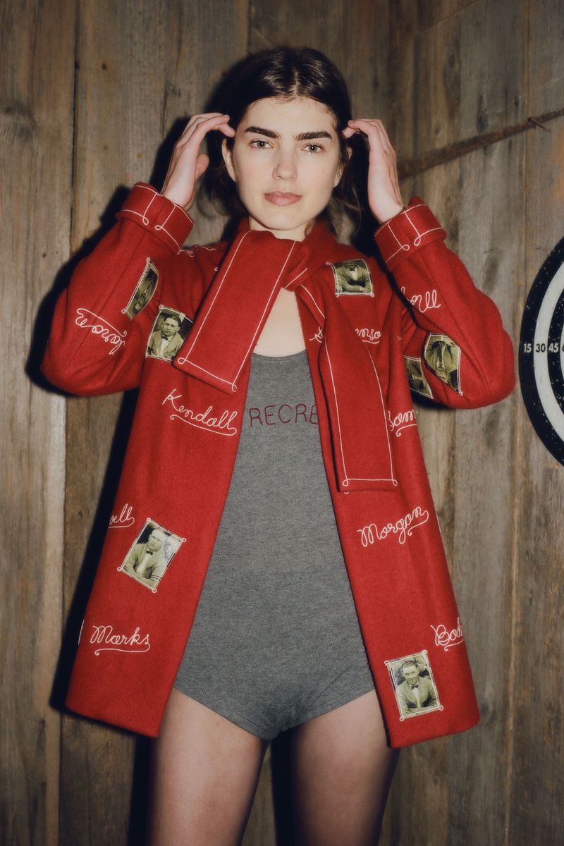 Woman in red jacket with photos poses against wood wall next to dart board. Jacket features personal photos and embroidered names.