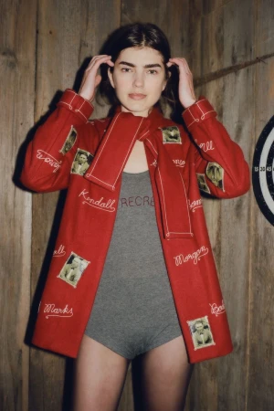 Woman in red jacket with photos poses against wood wall next to dart board. Jacket features personal photos and embroidered names.