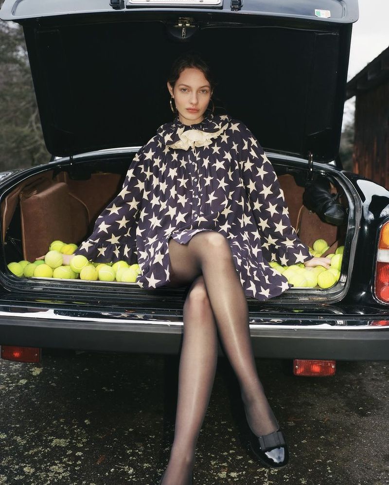 A woman models a stars-and-stripes poncho while seated in the open trunk of a car surrounded by tennis balls in an outdoor setting.