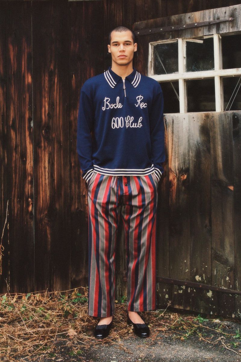 Full-shot fashion portrait of a slender adult male in a navy collared shirt with striped pants, posing against a weathered wooden wall outdoors.