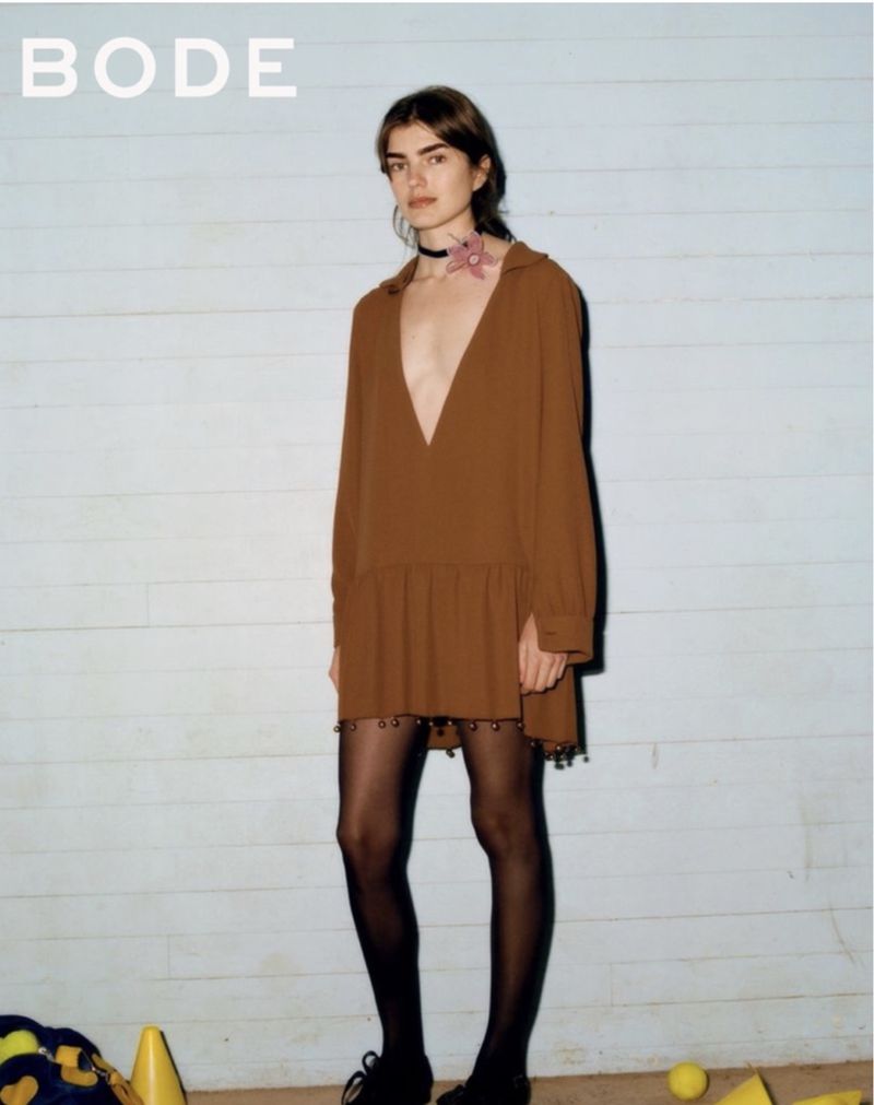 A woman poses in a brown dress, black tights, and black shoes, set against a pale blue wall and floor with scattered yellow objects.