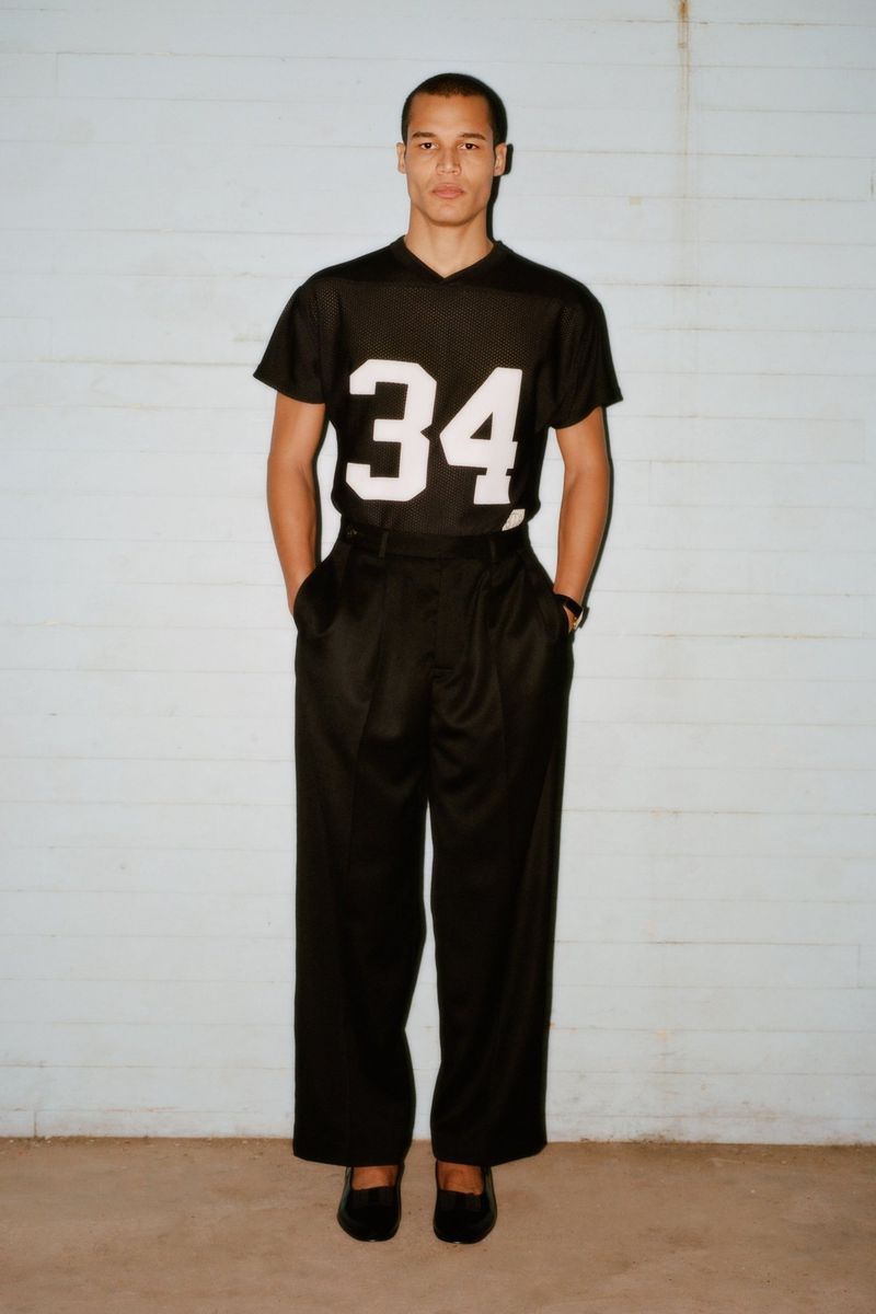 A male model poses in a black jersey with the number 34, wide black pants, and black shoes against a pale blue wall backdrop.