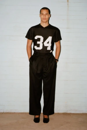 A male model poses in a black jersey with the number 34, wide black pants, and black shoes against a pale blue wall backdrop.