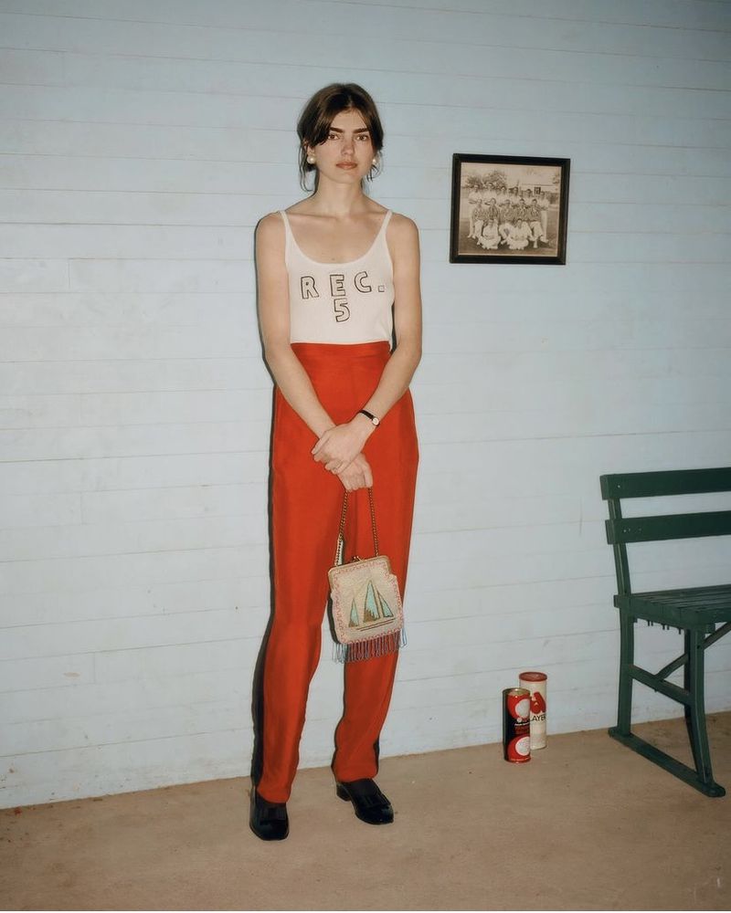 A young woman stands in front of a light blue wall wearing a white tank top and red pants, holding a beaded purse.