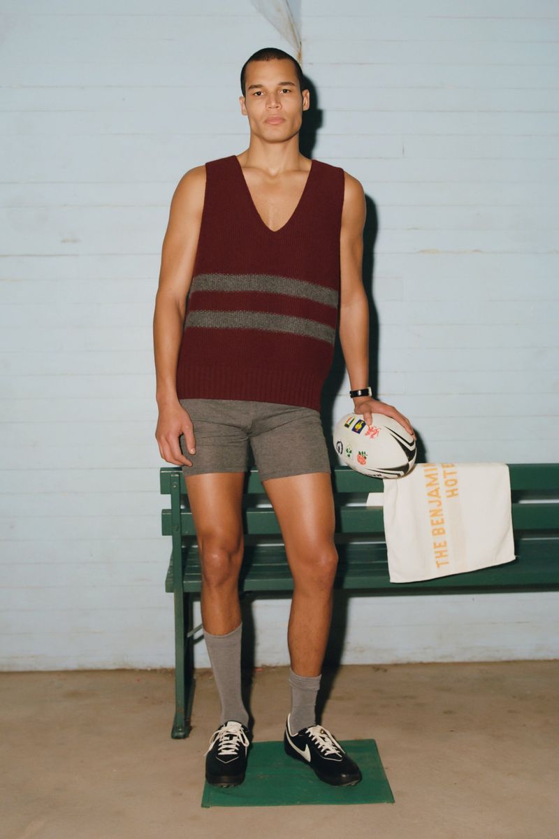 A man stands in a burgundy striped sweater vest and grey shorts, holding a football near a green bench in front of blue walls.