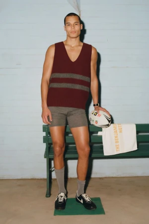 A man stands in a burgundy striped sweater vest and grey shorts, holding a football near a green bench in front of blue walls.
