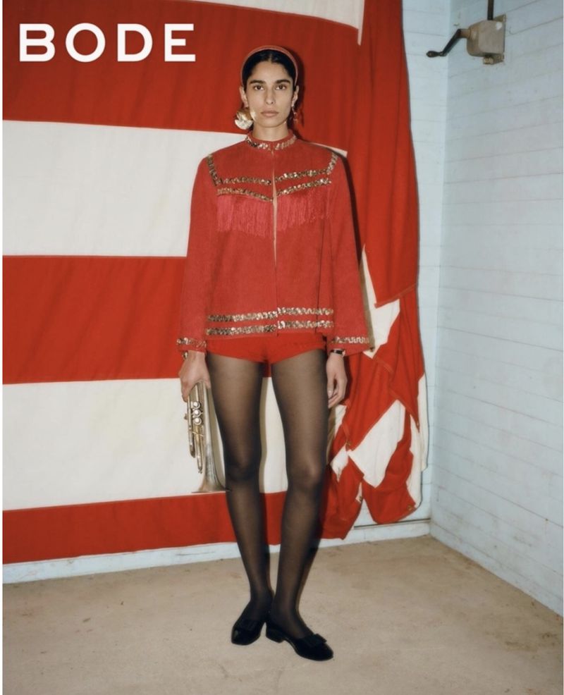 Adult female fashion model in red jacket, matching shorts, and black tights posing in front of a red-and-white striped curtain.