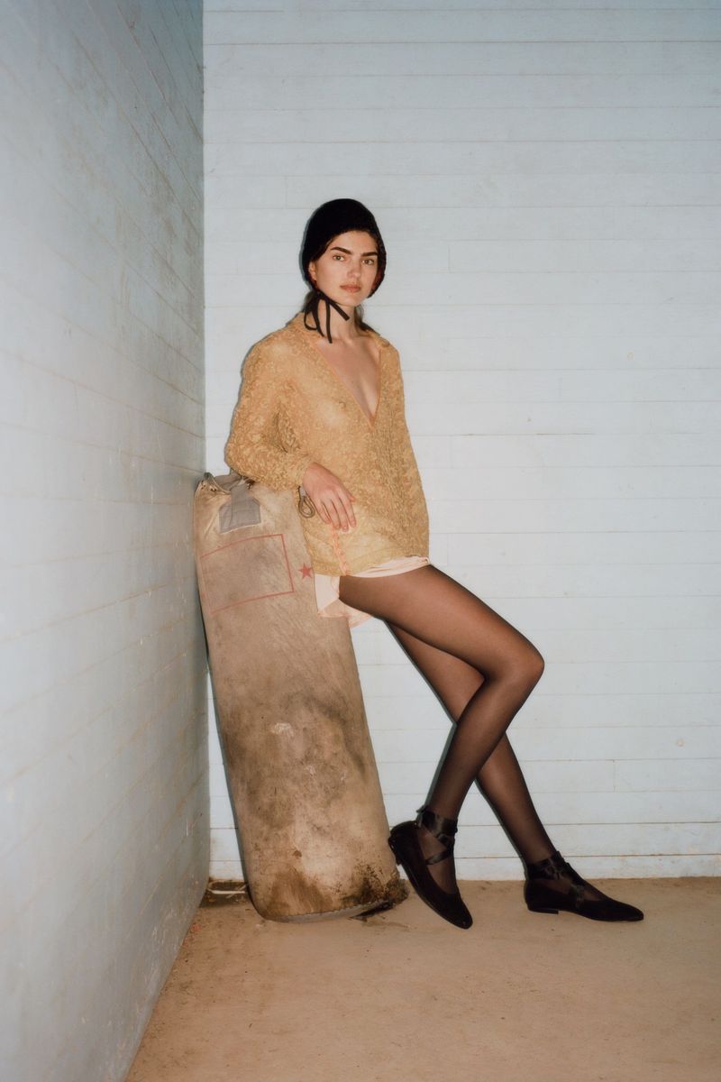 A stylish young woman with dark hair poses in a sheer shirt, mini shorts, dark tights, and flats, leaning on a large sack indoors.