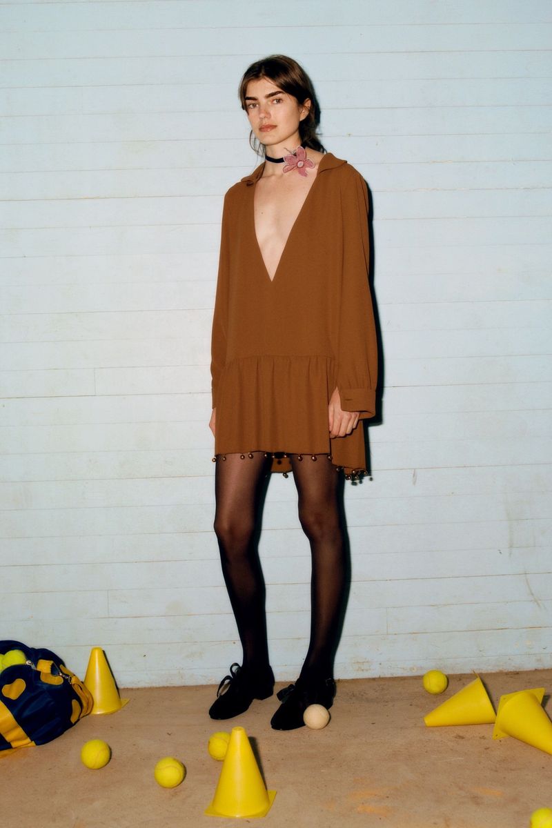 Woman in brown dress, black tights, and black shoes posing indoors against a light blue wall with yellow cones and balls scattered around.