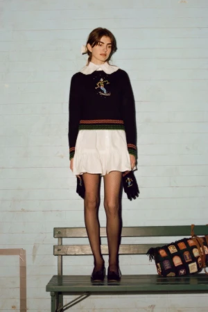 Young woman stands on a green bench with a knitted bag, wearing a sweater, white skirt, black tights, and black shoes in an outdoor setting.