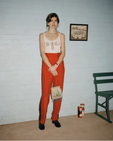 Model standing against a pale blue wood wall wearing a printed white tank top and red trousers holding a small embellished handbag