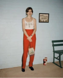 Model standing against a pale blue wood wall wearing a printed white tank top and red trousers holding a small embellished handbag