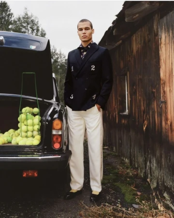 Model standing next to an open car trunk filled with tennis balls, wearing a dark numbered blazer and loose white trousers.