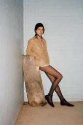 Model leaning against a heavy textured punching bag wearing a textured knit top, black tights, and shoes in a corner studio setting.