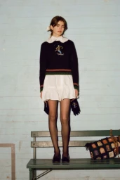 Model standing on a green wooden bench wearing a black embroidered knit sweater, white mini dress, and sheer black tights.