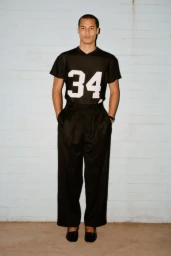 A male model stands in a black short-sleeve athletic jersey featuring number 34 tucked into black wide-leg trousers against a wall.