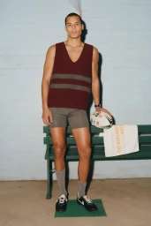 Model wearing a burgundy striped knit vest and grey shorts standing by a green bench holding a rugby ball.
