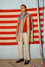 Model wearing an Americana-inspired fringe jacket and cream trousers standing before a large American flag backdrop for Bode.