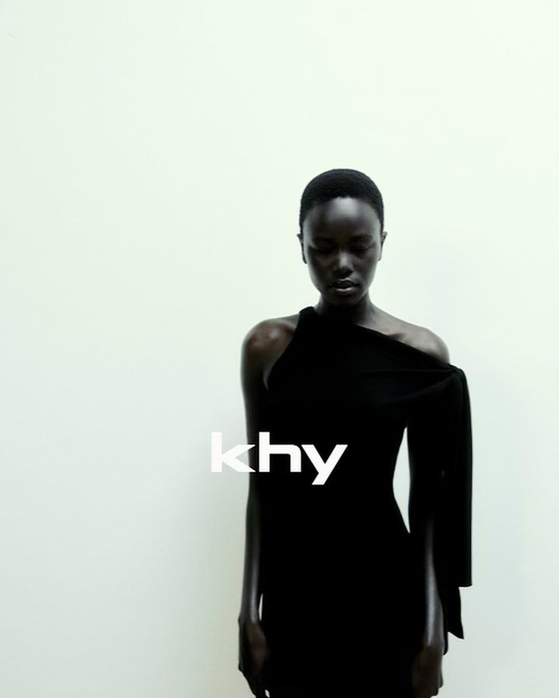 An African-American woman poses in a black dress against a stark white background, showcasing a high-fashion, minimalist aesthetic.