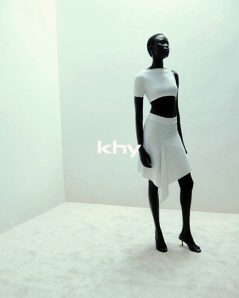 A slender, dark-skinned woman stands in a bright, minimalist studio wearing a white crop top, skirt, and high heels, showcased in a fashion shot.