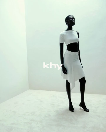 A slender, dark-skinned woman stands in a bright, minimalist studio wearing a white crop top, skirt, and high heels, showcased in a fashion shot.