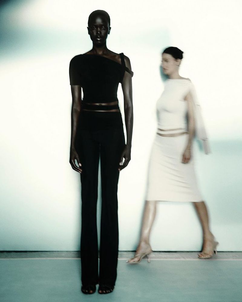 Two fashion models pose in a studio. One African-American woman in a black outfit stands, and the other woman is blurred in motion.