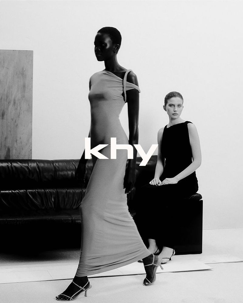 Two fashion models in a photo studio, one walking and one seated, wearing elegant evening gowns and heels, promoting the KHY brand.