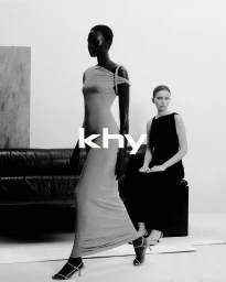 Two female models posing in a minimalist studio setting with a dark leather sofa and a plain white background.