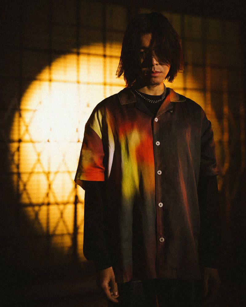 Portrait of a young man with long dark hair wearing a patterned button-down shirt, posing in front of a patterned wall with golden light.