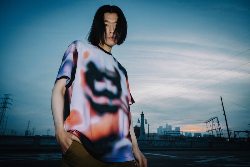 Adult man in a t-shirt poses on a street in front of a city skyline with a cloudy sky during the day.
