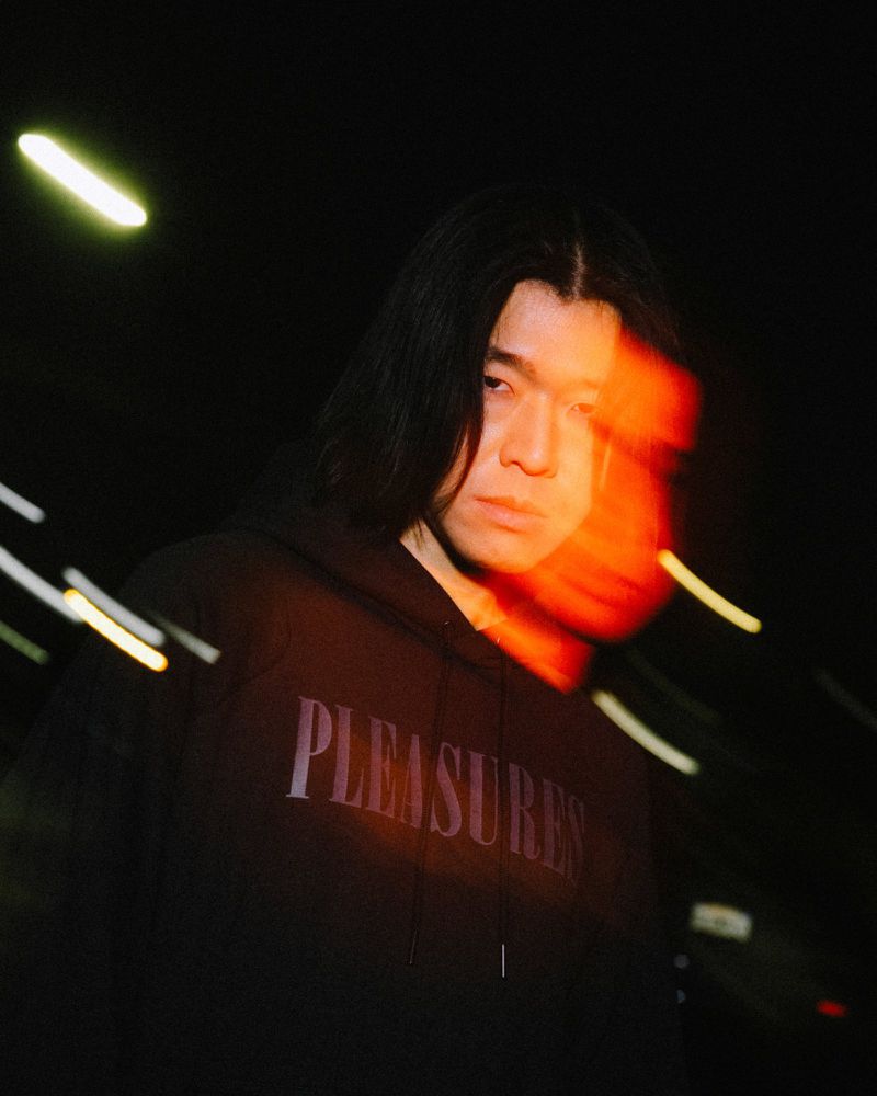 Portrait of an adult male wearing a black hoodie with the word 'PLEASURES' in front, posing in a dark outdoor environment.