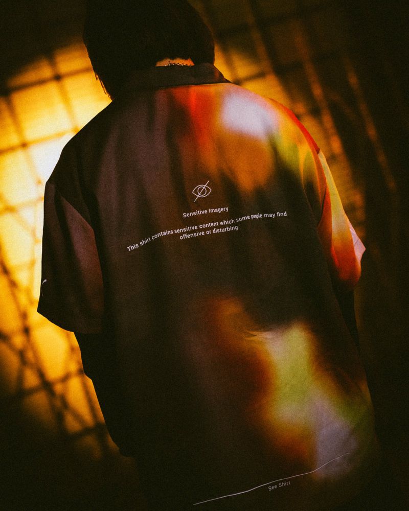 Person wears an artistic button-down shirt with light and shadow play. The shirt contains a sensitive imagery message against a warm background.